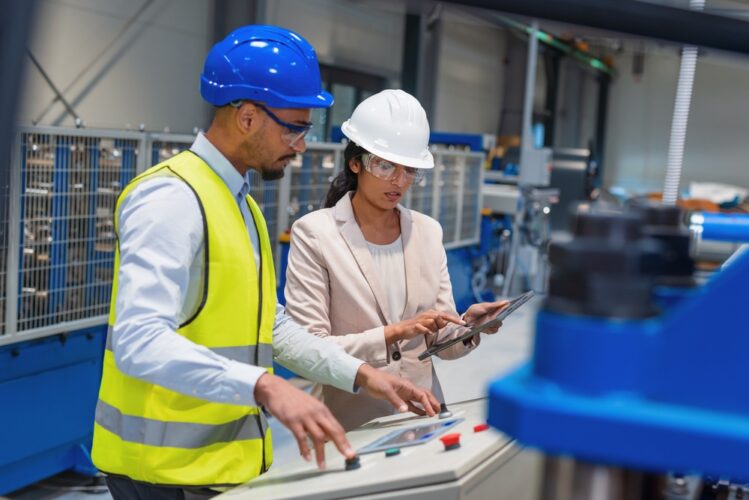 An engineer and a technician in a factory setting, both wearing hard hats and safety glasses. The engineer uses a tablet while the technician operates a control panel on a piece of large blue industrial machinery.