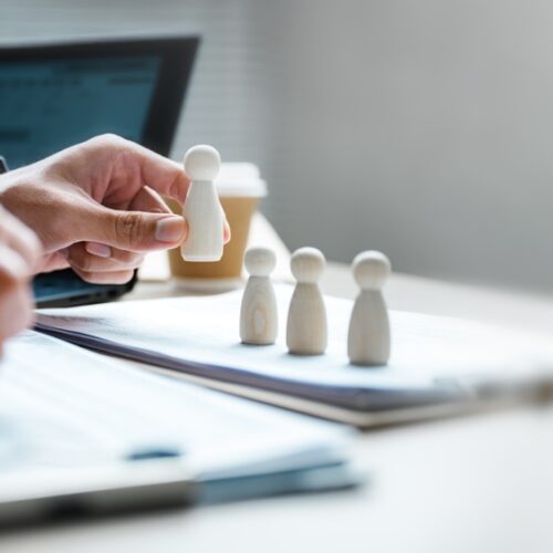 A close-up of a person's hand placing a wooden peg figure in a row with three others on a desk. The person is simultaneously writing on a document with their other hand, with a laptop and coffee cup visible in the blurred background, symbolizing recruitment or human resources management.