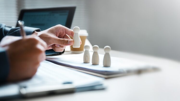 A close-up of a person's hand placing a wooden peg figure in a row with three others on a desk. The person is simultaneously writing on a document with their other hand, with a laptop and coffee cup visible in the blurred background, symbolizing recruitment or human resources management.