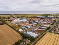 An aerial view of an industrial area near the sea, featuring various warehouses, outdoor storage yards for heavy equipment, and adjacent residential streets and fields.