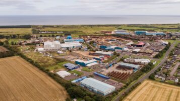 An aerial view of an industrial area near the sea, featuring various warehouses, outdoor storage yards for heavy equipment, and adjacent residential streets and fields.