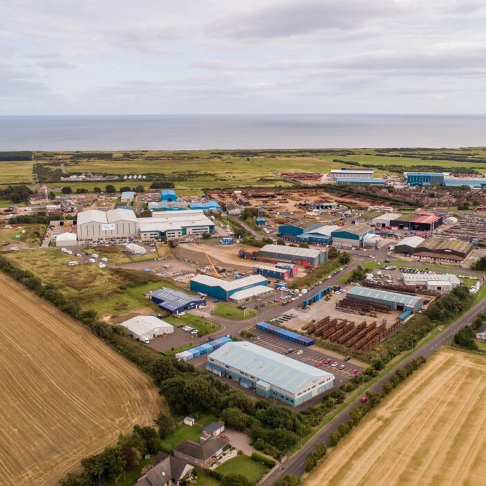 An aerial view of an industrial area near the sea, featuring various warehouses, outdoor storage yards for heavy equipment, and adjacent residential streets and fields.