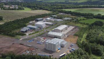 An aerial view of an industrial estate surrounded by green trees and fields. Several large metal-clad warehouses and storage yards are distributed throughout the park.
