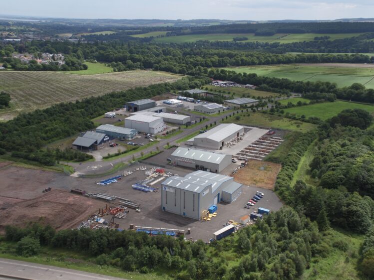 An aerial view of an industrial estate surrounded by green trees and fields. Several large metal-clad warehouses and storage yards are distributed throughout the park.