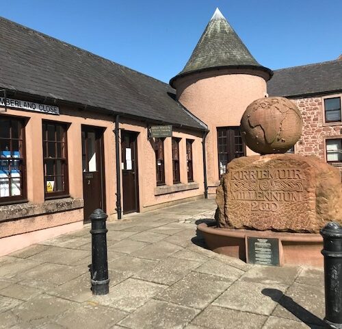 A stone building with a small turret and a sign reading "Cumberland Close." In the foreground, there is a large, rounded stone monument topped with a stone globe, engraved with "Kirriemuir Millennium 2000."