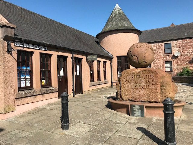 A stone building with a small turret and a sign reading "Cumberland Close." In the foreground, there is a large, rounded stone monument topped with a stone globe, engraved with "Kirriemuir Millennium 2000."