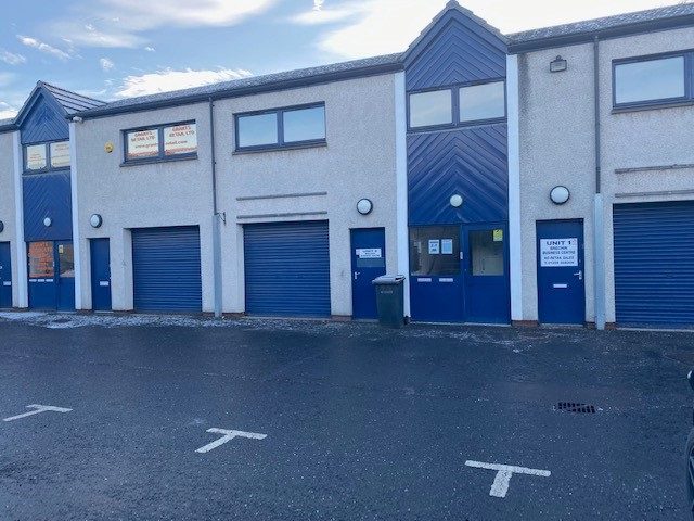 A row of light-colored industrial units with blue garage-style roller doors and matching blue trim around the pedestrian entrances. The units are located on a dark asphalt lot with white painted "T" parking markers.