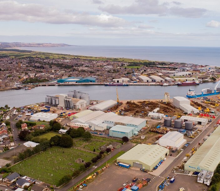 A high-angle panoramic view of a coastal town showing a dense industrial port area with large storage tanks and long warehouses, bordered by a cemetery and residential housing.