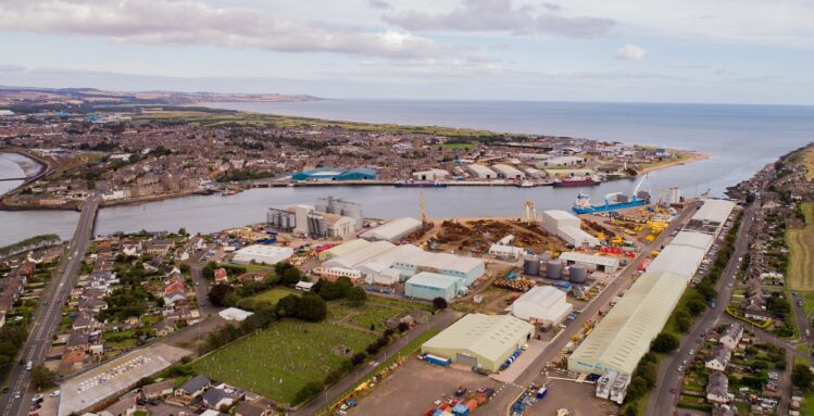 A high-angle panoramic view of a coastal town showing a dense industrial port area with large storage tanks and long warehouses, bordered by a cemetery and residential housing.
