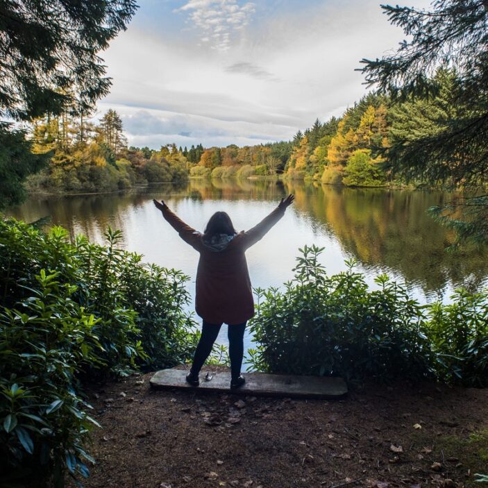 A person stands with arms outstretched facing a calm lake surrounded by dense evergreen and autumnal trees. The forest framing creates a natural window looking out over the water.