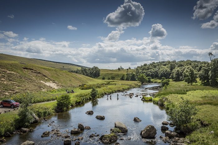 A wide landscape of a river winding through a lush green valley under a bright, cloudy sky. People are wading in the shallow water, and a red car is parked on the grassy bank to the left.
