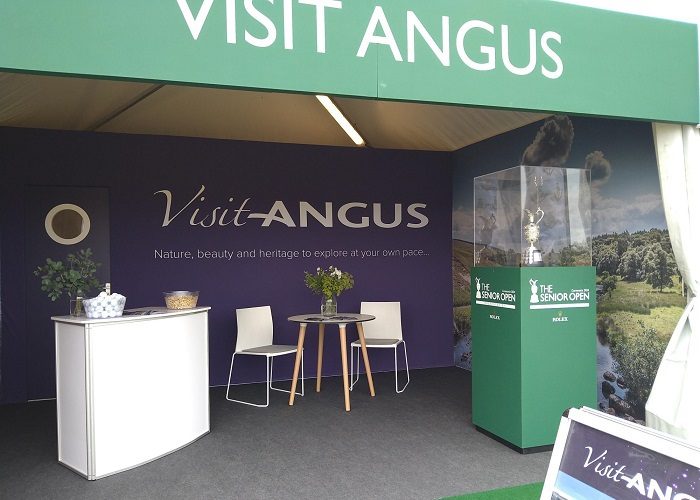 An exhibition stand for "Visit Angus" at the 2024 Senior Open golf tournament. The booth features purple backdrops with the slogan "Nature, beauty and heritage to explore at your own pace," a white reception counter, and a small table with two chairs. To the right, the Senior Open trophy is displayed in a glass case on a green pedestal.