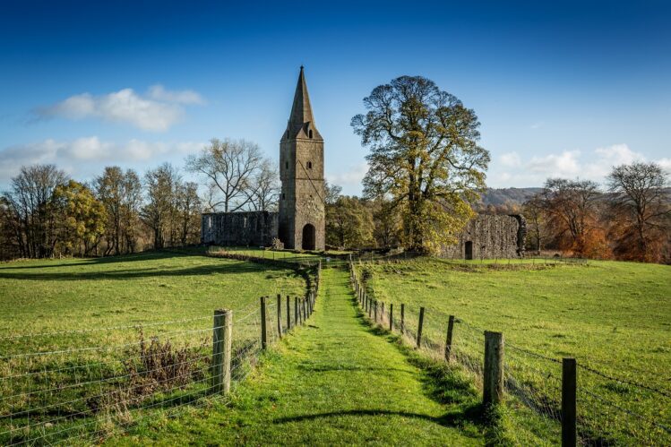 A wide landscape shot of Restenneth Priory in Angus, Scotland, under a clear blue sky. A narrow grass path, flanked by wire fences, leads through a bright green field toward the ancient stone ruins. The most prominent feature is the priory's tall, slender square tower topped with a stone spire, surrounded by autumnal trees and rolling hills in the background.