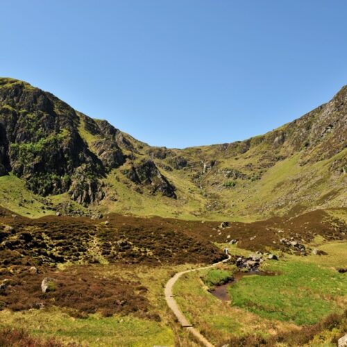 A sweeping landscape view of Corrie Fee National Nature Reserve in the Angus Glens. A narrow dirt path winds through a floor of brown heather and green grass, leading toward a massive, glacier-carved amphitheater of steep, rocky mountains under a clear blue sky. A small stream meanders alongside the trail in the foreground.