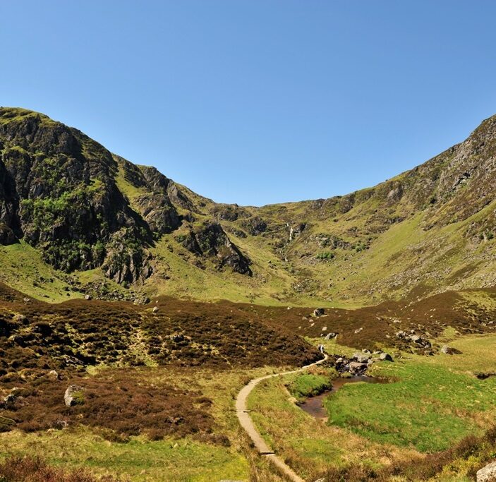 A sweeping landscape view of Corrie Fee National Nature Reserve in the Angus Glens. A narrow dirt path winds through a floor of brown heather and green grass, leading toward a massive, glacier-carved amphitheater of steep, rocky mountains under a clear blue sky. A small stream meanders alongside the trail in the foreground.