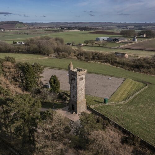 An aerial drone photograph of the Balmashanner War Memorial (locally known as "Bummy") in Forfar, Angus. The tall, square stone tower features a battlement top and a smaller circular turret. It stands on a hill overlooking a patchwork of green and brown farmland, with farm buildings and clusters of trees scattered across the wide Scottish landscape under a clear sky.