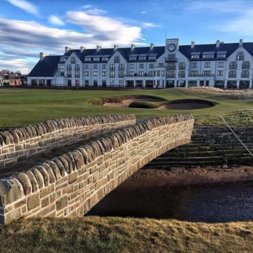 A stone bridge with rounded masonry walls crosses a small burn in the foreground, leading toward the green fairways, sand bunkers, and the large white facade of the Carnoustie Golf Hotel under a bright, partly cloudy sky.