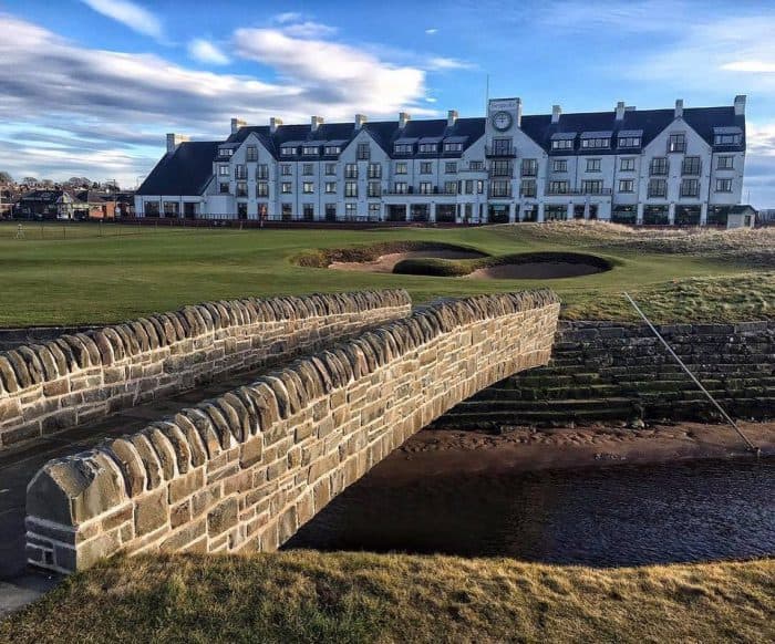 A stone bridge with rounded masonry walls crosses a small burn in the foreground, leading toward the green fairways, sand bunkers, and the large white facade of the Carnoustie Golf Hotel under a bright, partly cloudy sky.