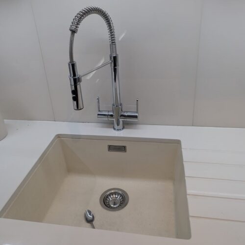 A modern chrome kitchen tap with a high-arched spring neck and pull-down sprayer, set above a beige undermount sink with a single spoon inside.