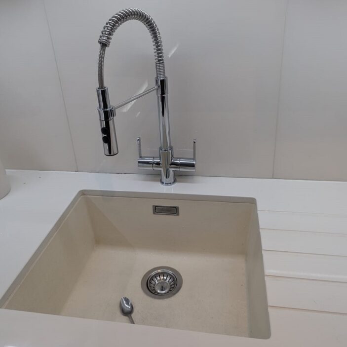 A modern chrome kitchen tap with a high-arched spring neck and pull-down sprayer, set above a beige undermount sink with a single spoon inside.