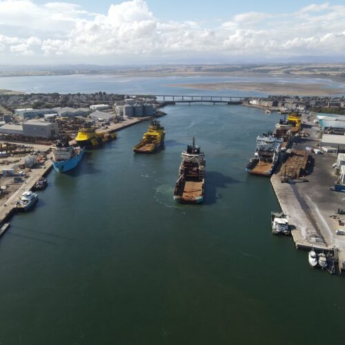 An aerial view of a busy harbor with several large industrial and offshore supply vessels docked at piers or anchored in the channel, leading toward a bridge and a coastal town under a bright, cloud-filled sky.