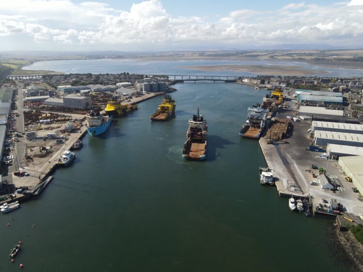An aerial view of a busy harbor with several large industrial and offshore supply vessels docked at piers or anchored in the channel, leading toward a bridge and a coastal town under a bright, cloud-filled sky.