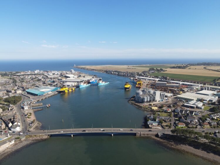 A high-angle aerial view of a busy port and coastal town. Several large industrial ships, including yellow and blue vessels, are docked or moving through the deep blue channel. A bridge with cars crossing spans the water in the foreground, leading toward the sea and a distant lighthouse.