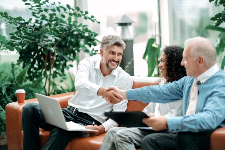 Three people sit on an orange sofa in a bright, modern office lobby filled with greenery. A middle-aged man in a white shirt shakes hands with an older man in a blue shirt, while a woman with curly hair watches and smiles. A laptop rests on the lap of the man in the white shirt.