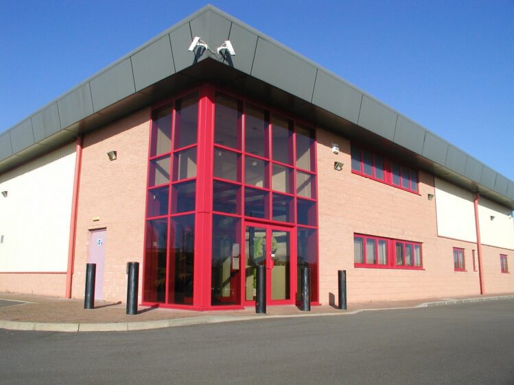 A modern two-story industrial office building at Brechin Business Park with a prominent red-framed glass entrance, tan brick walls, and a dark grey overhanging roofline under a clear blue sky.