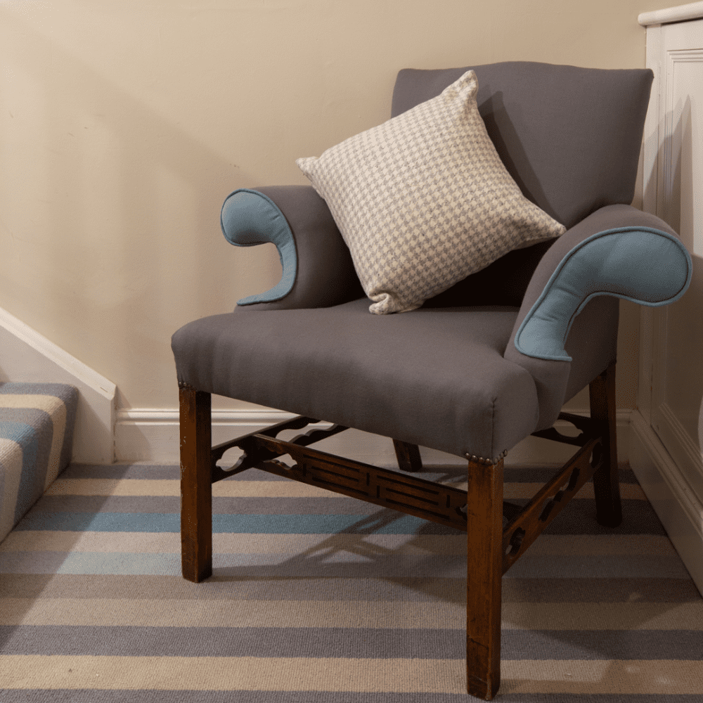 Marana Collection Hero Shoot - a grey upholstered armchair with light blue rolled arm accents sits on a striped carpet in soft neutral tones. A small patterned cushion is placed on the seat, and the chair stands beside a staircase and a white wall.