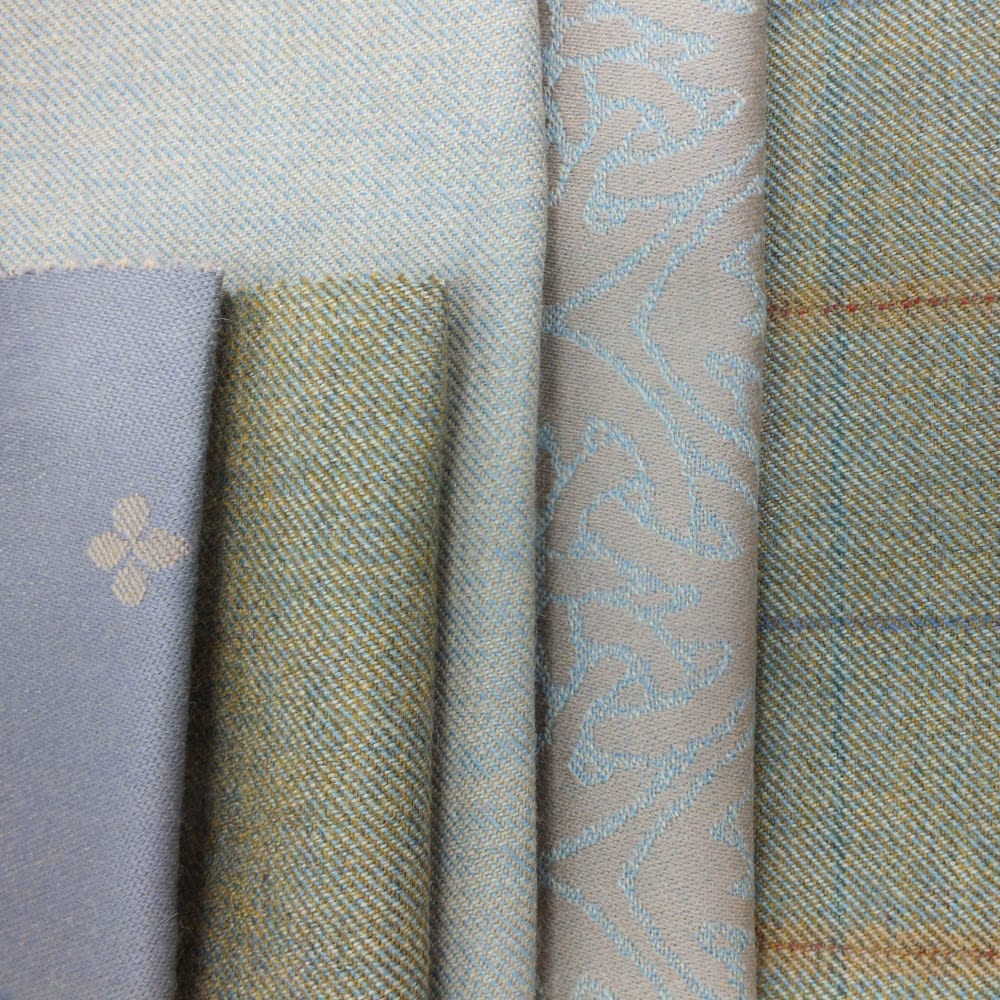 Clunie Burn collection hero photo - selection of folded fabrics in soft blue and muted green tones, featuring a mix of textures and patterns, including herringbone, jacquard, and subtle checks.