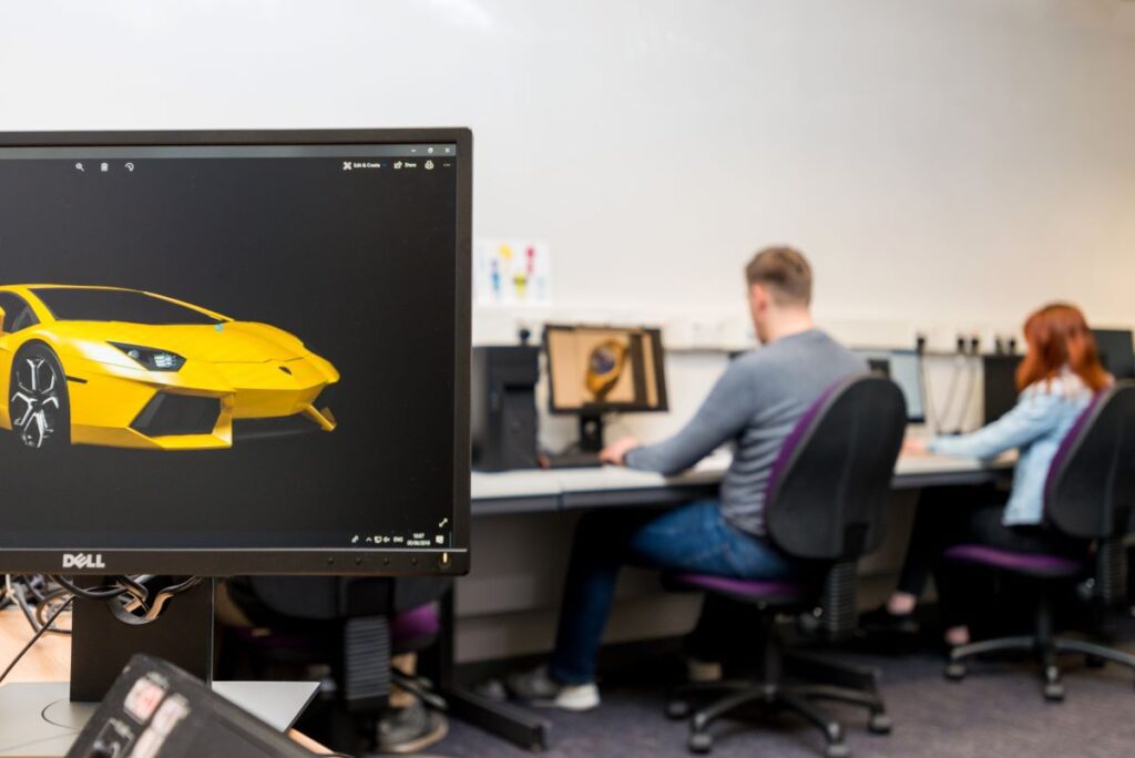 A computer screen with a yellow sports car on it. In the background, two students are working at computers.