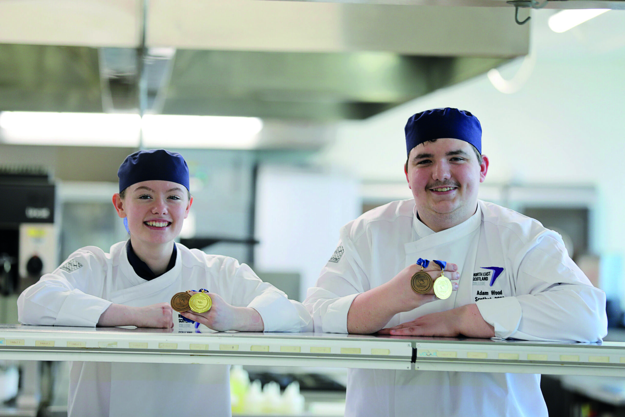 Adam and Ellie, two hospitality students, stood in the NESCol kitchens, holding medals and smiling.