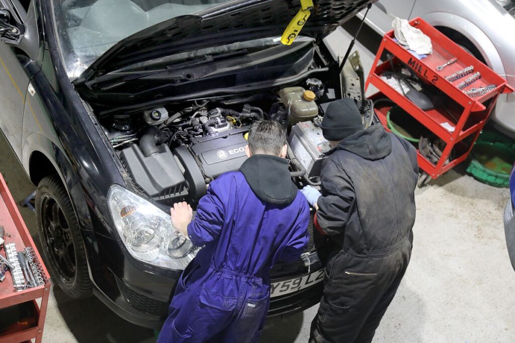 Two students in boilersuits working under the hood of a car.