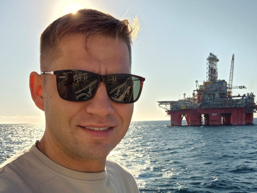 Headshot of Andrius offshore wearing sunglasses. In the background is the sea and an oil rig.