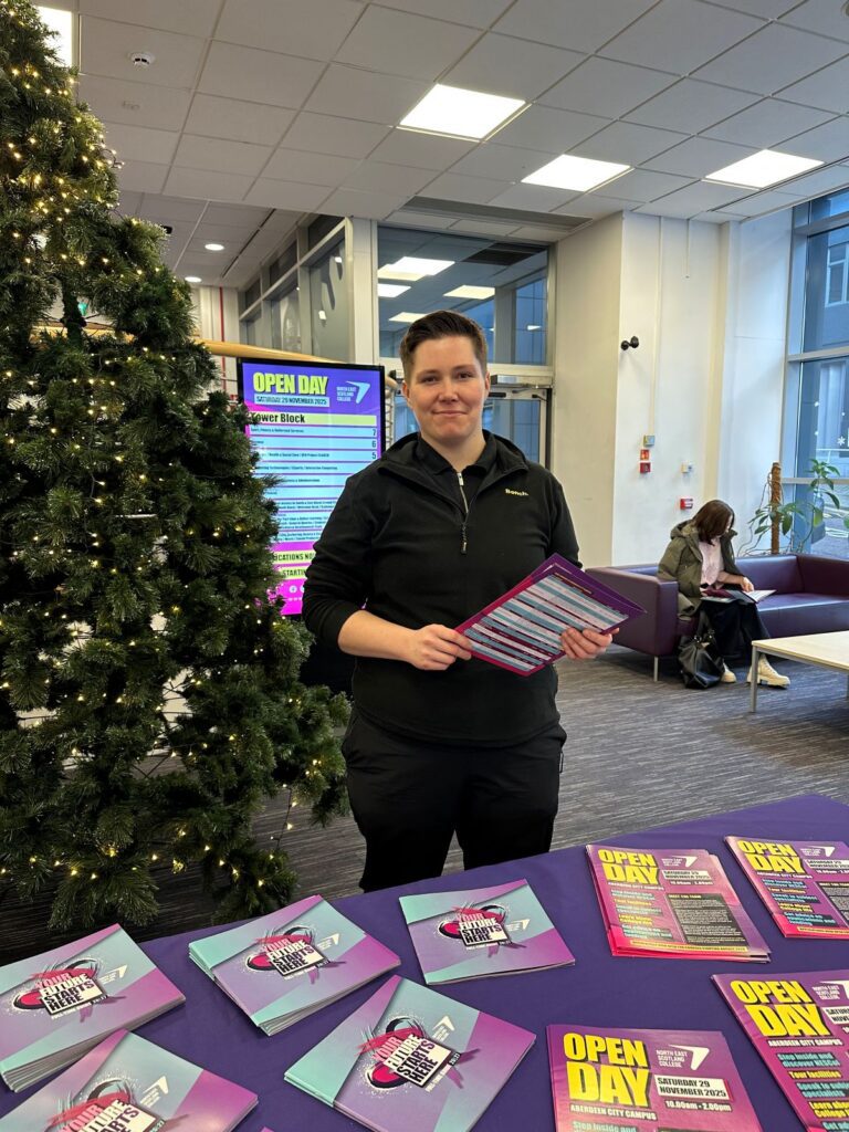 Weronika at the NESCol Open day standing in front of a table of flyers and next to a Christmas tree.