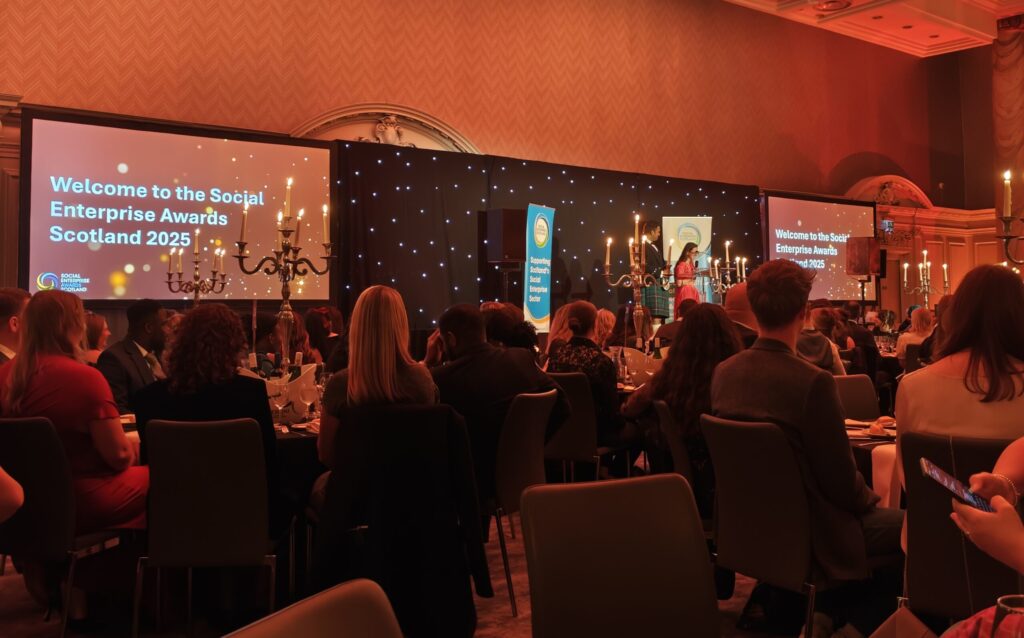 A photo taken at the 2025 Social Enterprise Scotland Awards. Groups of people sit around tables in a ballroom and watch someone receive an award on stage