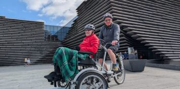 A photo of 2 men, one on a bike and one on a wheelchair attached to the front of the bike. they are posing in front of V&A Dundee