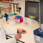 David Gray, wearing lab coat and safety glasses, sits at a lab bench.