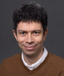 Head shot of Manu. He has short dark hair. He is wearing a white shirt under a brown jumper.