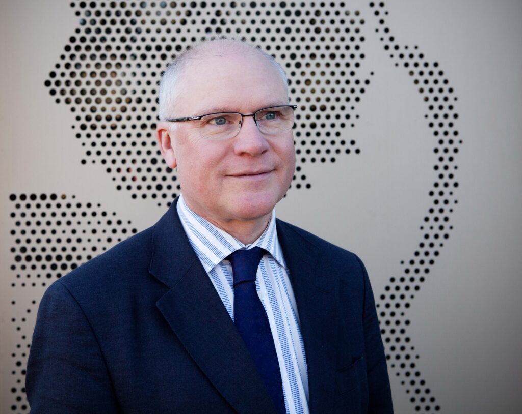 Head and shoulders shot of Mike Ferguson standing in front of one of the panels of the Discovery building. Mike is wearing a dark suit and tie with a white shirt.
