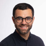 Douglas has short dark hair, a short beard and wears round black rimmed glasses