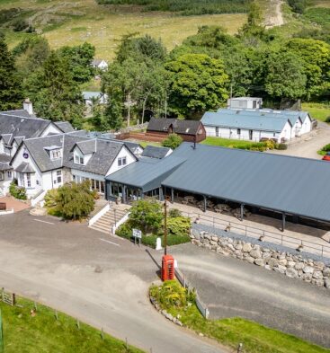 An elevated wide shot of the Glen Clova Hotel, showing a white multi-story building with dark grey roofs, a modern restaurant extension, and a red telephone box surrounded by green hills.