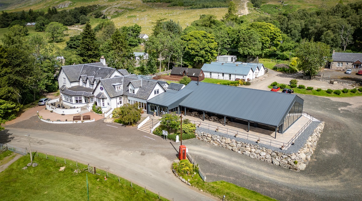 An elevated wide shot of the Glen Clova Hotel, showing a white multi-story building with dark grey roofs, a modern restaurant extension, and a red telephone box surrounded by green hills.