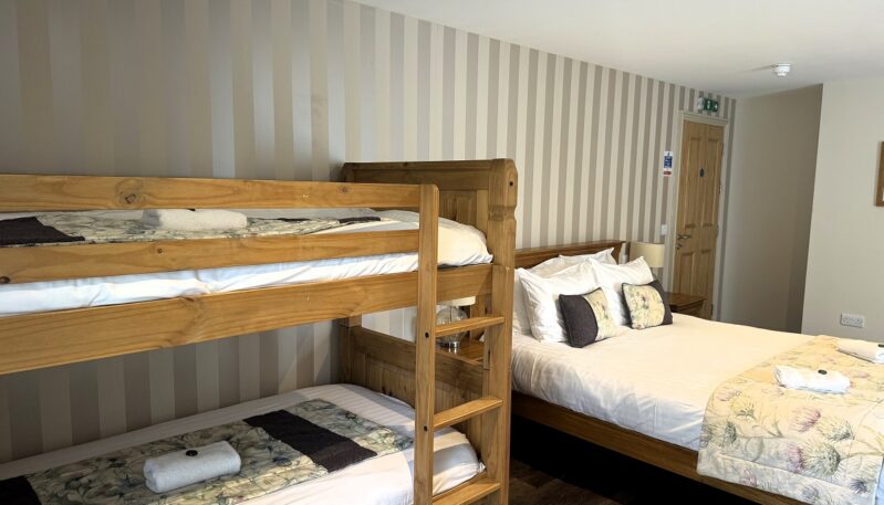 A rustic hotel family room featuring a set of wooden bunk beds in the foreground and a large double bed in the background, set against a neutral striped wallpaper.