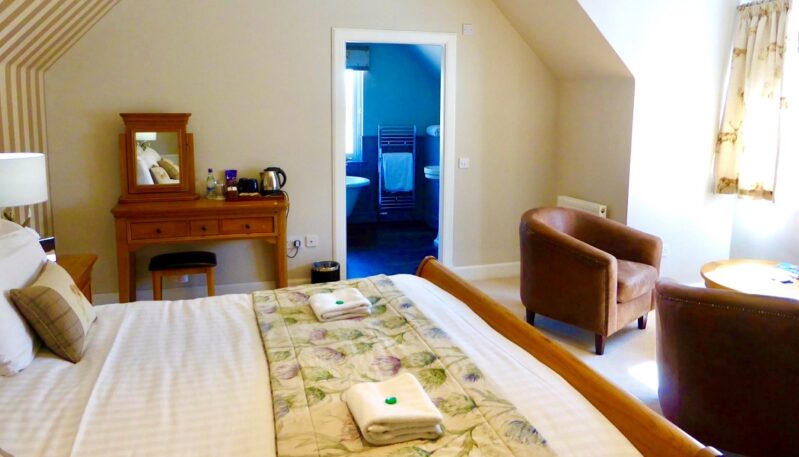A bright, cozy junior suite with a wooden bed frame, leather armchairs, a classic dressing table, and an open door leading to a modern bathroom with a blue accent wall.