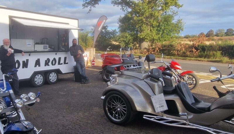 A wide outdoor shot featuring a white food trailer labeled "ON A ROLL" on the left, with two men standing near the serving window. Several Rewaco trikes (three-wheeled motorcycles) are parked in the foreground and middle ground. The most prominent trike is a light brown/tan model with a price tag of $\textsterling 18,995$ on its fender, with a red trike and a blue trike also visible. A Rewaco flag is positioned behind the food van. The scene is set in a paved area next to a road, with trees and countryside visible in the background.