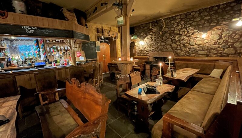 A traditional, dimly lit bar with a stone feature wall, rustic live-edge wooden tables and benches, and a wooden bar counter lit by warm candlelight.