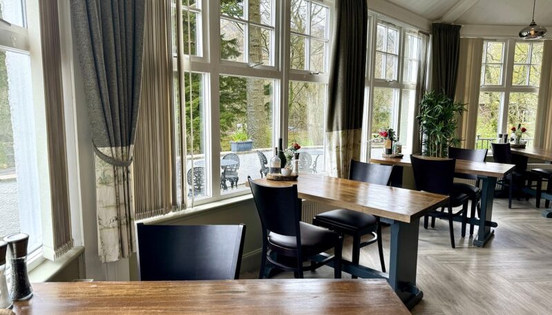 A light-filled contemporary restaurant with large windows overlooking a patio, featuring simple wooden tables, dark chairs, and light herringbone-patterned flooring.