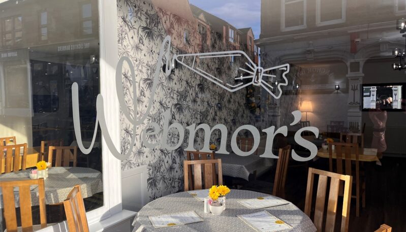 A view through a window with the Webmor's logo, showing an interior dining area with round tables, yellow flowers, and patterned wallpaper.